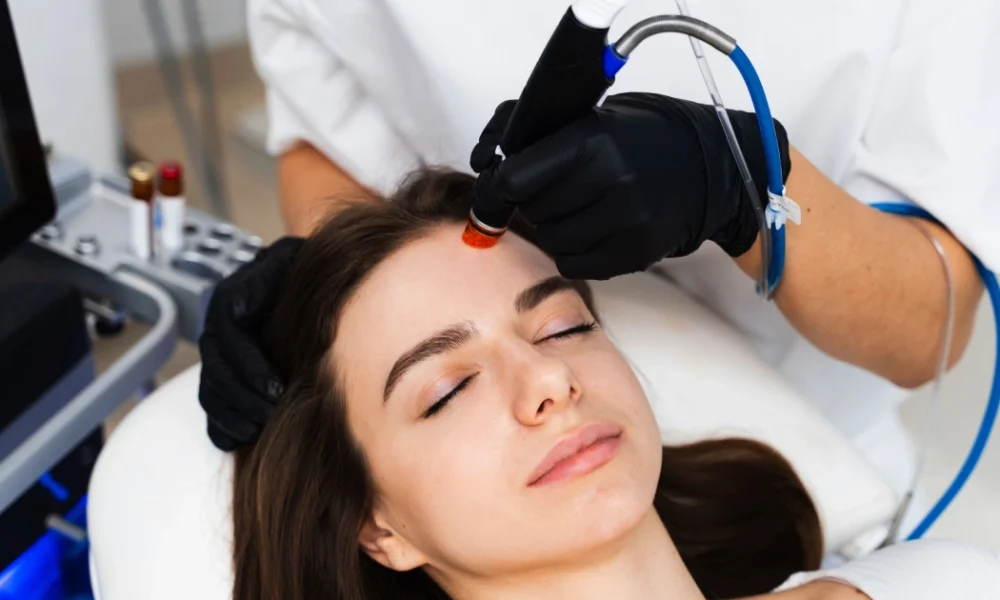 A woman receiving a HydraFacial in Houston with a handheld device from a person wearing black gloves.